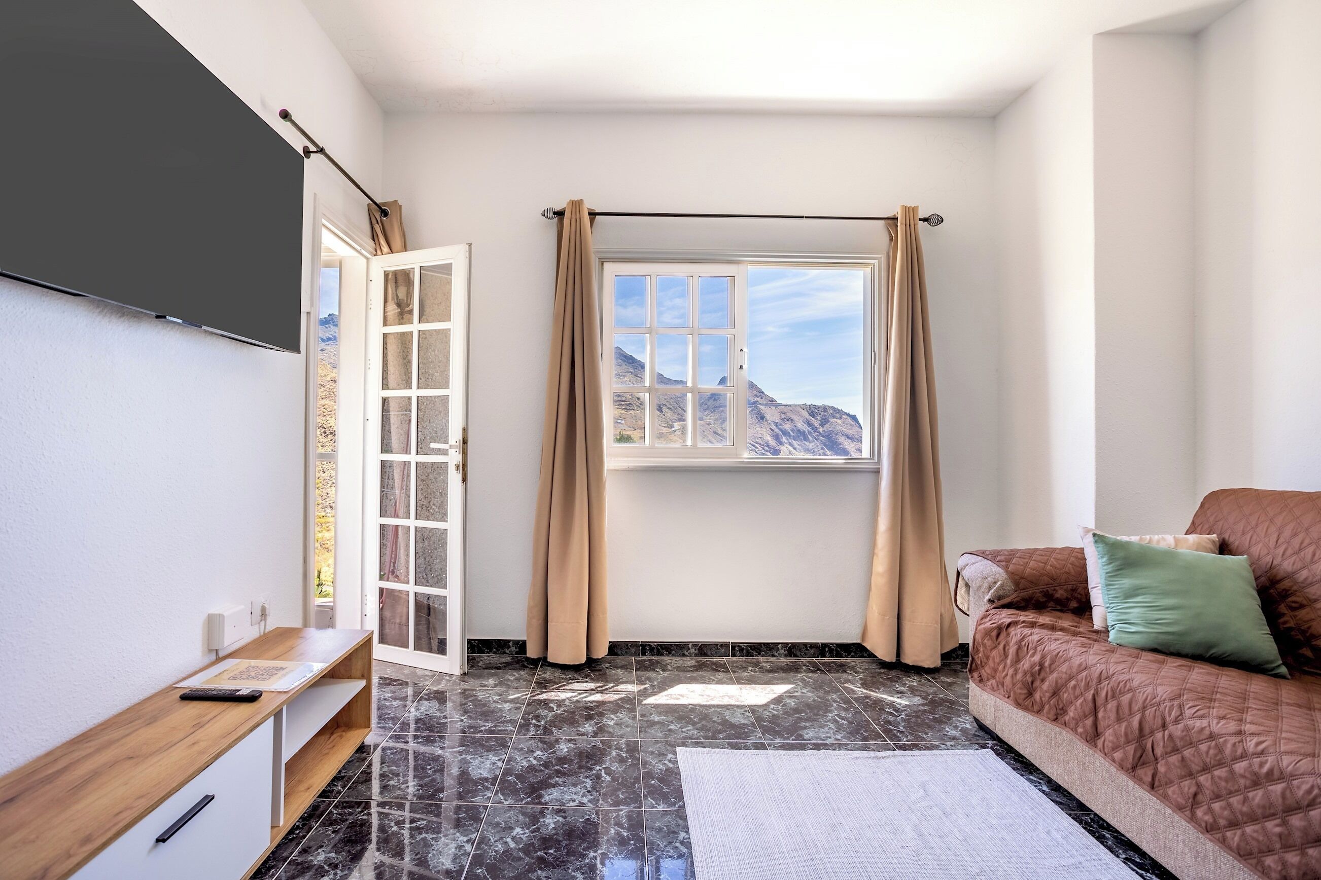 A room with a bed, a TV, a window with curtains, a door, and a view of mountains.