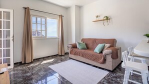 Um quarto com um sofá bege, uma mesa de jantar com cadeiras, uma janela com cortinas e uma estante com plantas.