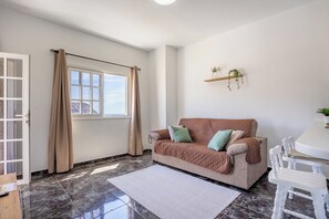 Ein Zimmer mit einem beigefarbenen Sofa, einem Esstisch mit Stühlen, einem Fenster mit Vorhängen und einem Regal mit Pflanzen.