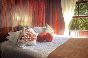 Una cama con colcha blanca y almohadones decorativos, una pared de madera y una ventana con vistas a la vegetación.