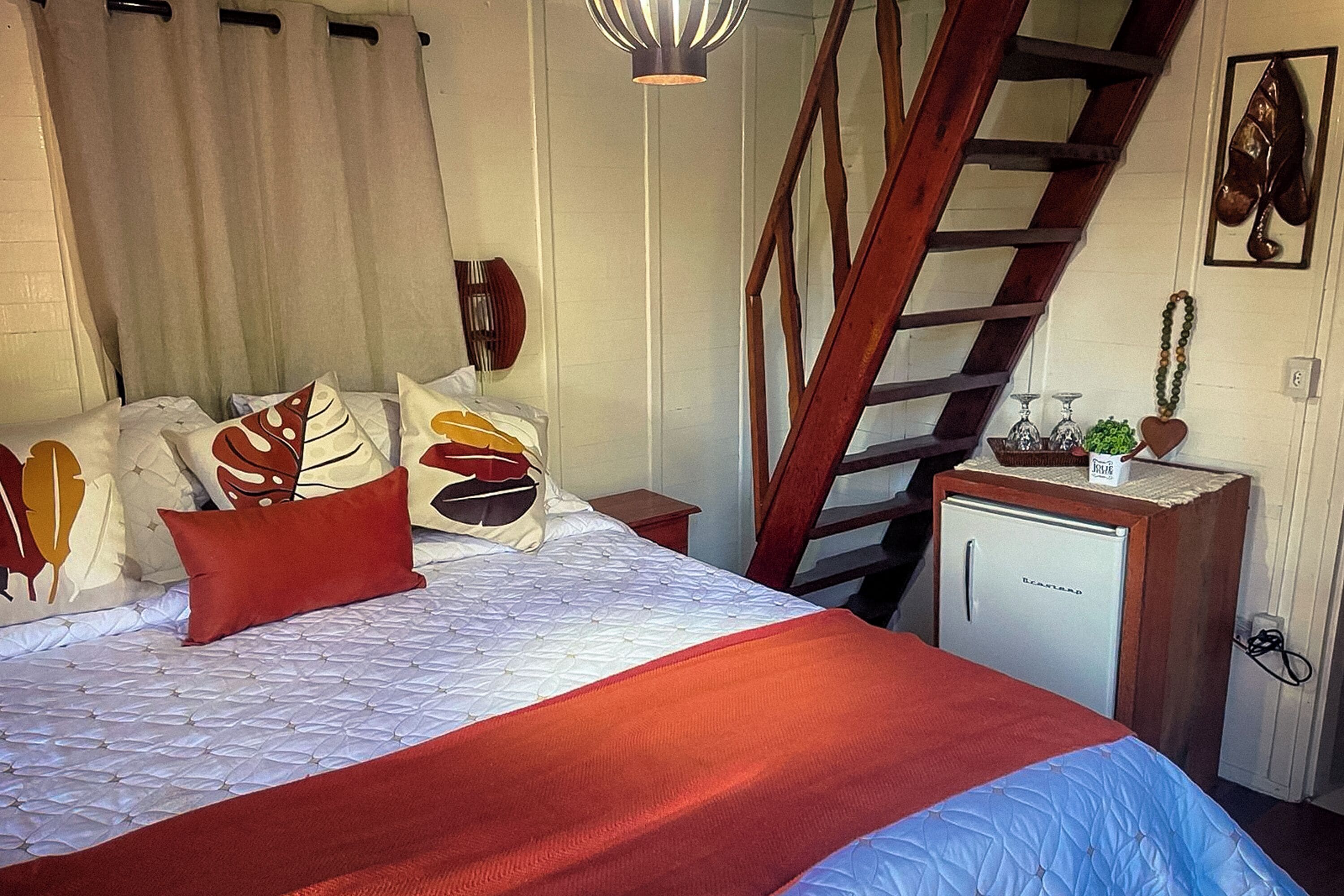 Un dormitorio con cama, almohadas, mesita de noche, cómoda, escalera y un refrigerador pequeño.