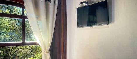 A room with a window, white curtains, and a mounted television on the wall.
