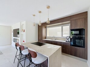 A modern kitchen with a central island, bar stools, wooden cabinets, and a window above the sink. - Villa Neuve avec Jardin Tropical à 700m de la mer (AGDE)