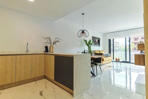 A modern kitchen with a marble countertop, wooden cabinets, a dining area with a sofa and table, and a large window offering a view of the outside. - Groundfloor Villamartin Amanecer (Orihuela)