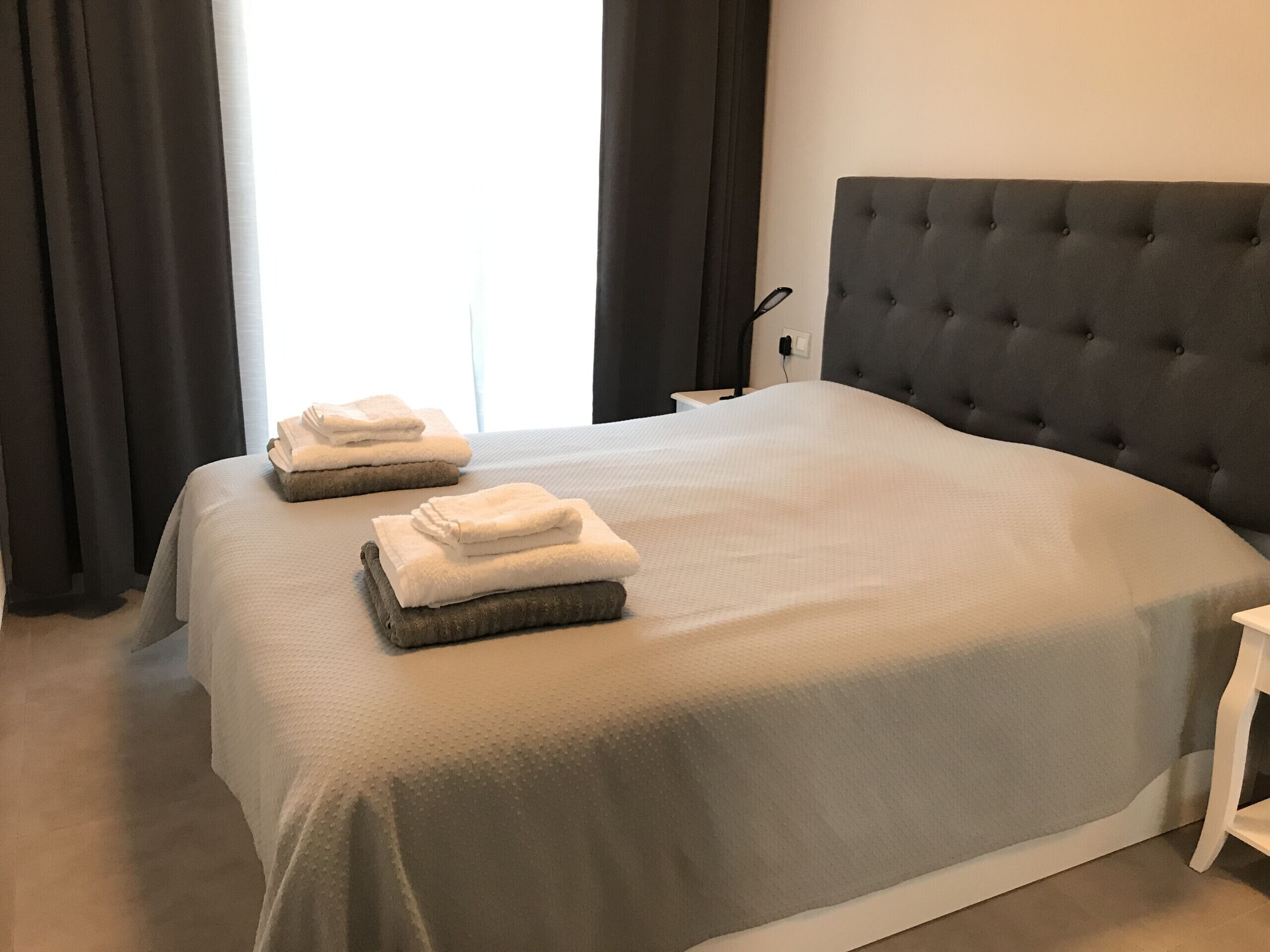 Een netjes opgemaakt bed met een grijze hoofdbord en een witte sprei. Er liggen twee opgevouwen handdoeken op het bed.