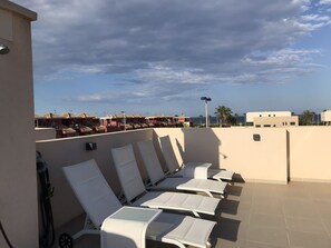 Eine Dachterrasse mit weißen Liegestühlen und Blick auf das Meer sowie Gebäude im Hintergrund.