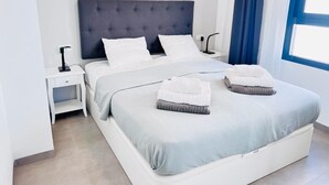 A modern bedroom with a large bed, white bedding, a dark headboard, a bedside table with a lamp, and a window with blue curtains. - Penthouse Higuericas Playas (Pilar de la Horadada)