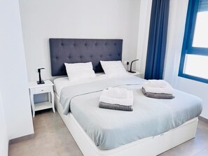 Ein modernes Schlafzimmer mit einem großen Bett, weißer Bettwäsche, einem dunklen Kopfteil, einem Nachttisch mit einer Lampe und einem Fenster mit blauen Vorhängen.
