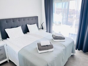 Habitación de hotel con una cama grande, dos almohadas, una mesita de noche con lámpara y vistas a la ciudad desde la ventana.