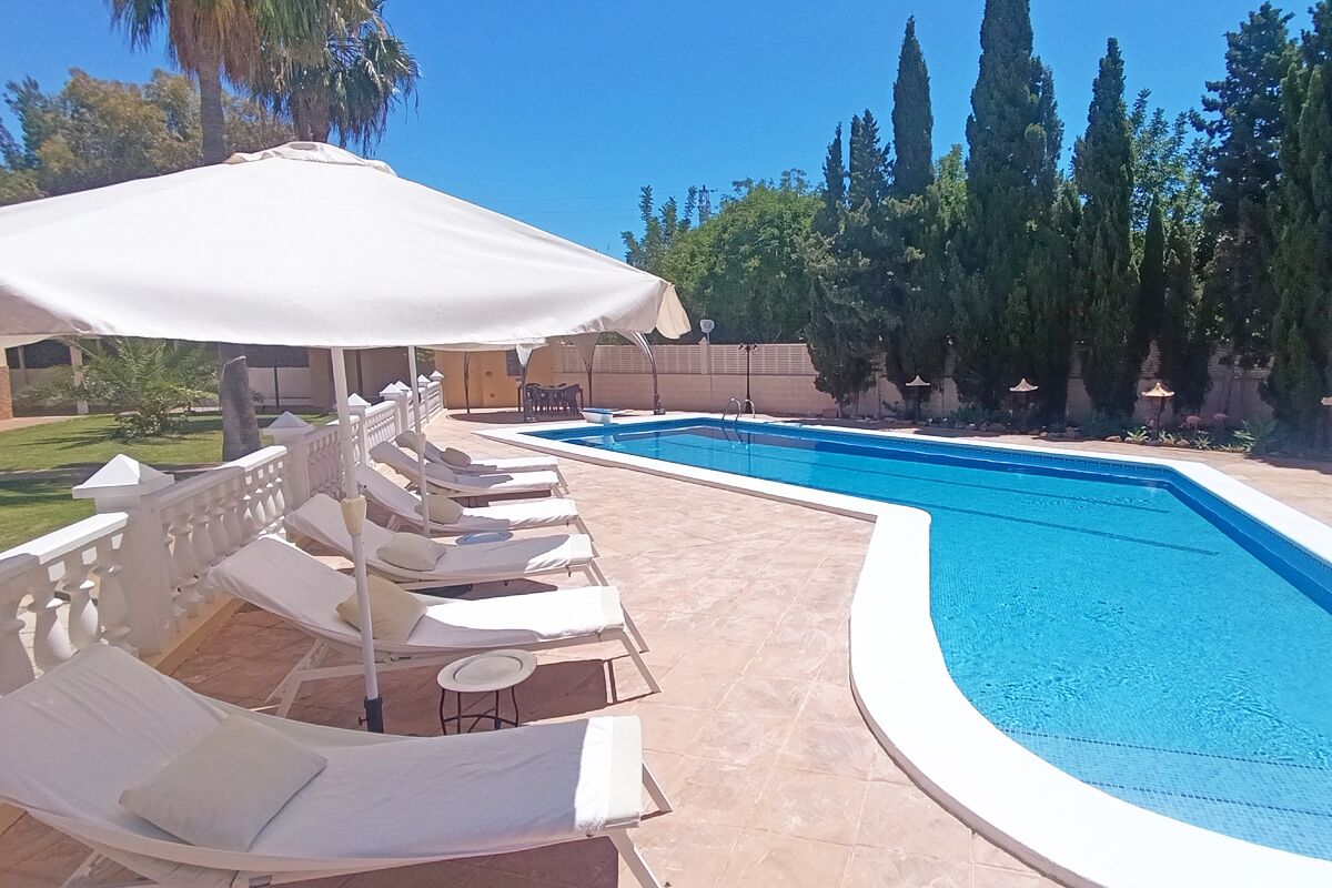 Une piscine entourée d’un jardin et de grands arbres, avec des chaises longues et des parasols à proximité.