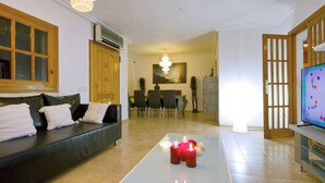 Uma sala de estar com sofá, mesa de centro com velas, televisão e um espaço de refeições ao fundo.