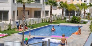 A swimming pool area with a pool, palm trees, and a fenced area. - Groundfloor Property Amalia Torrevieja (Torrevieja)