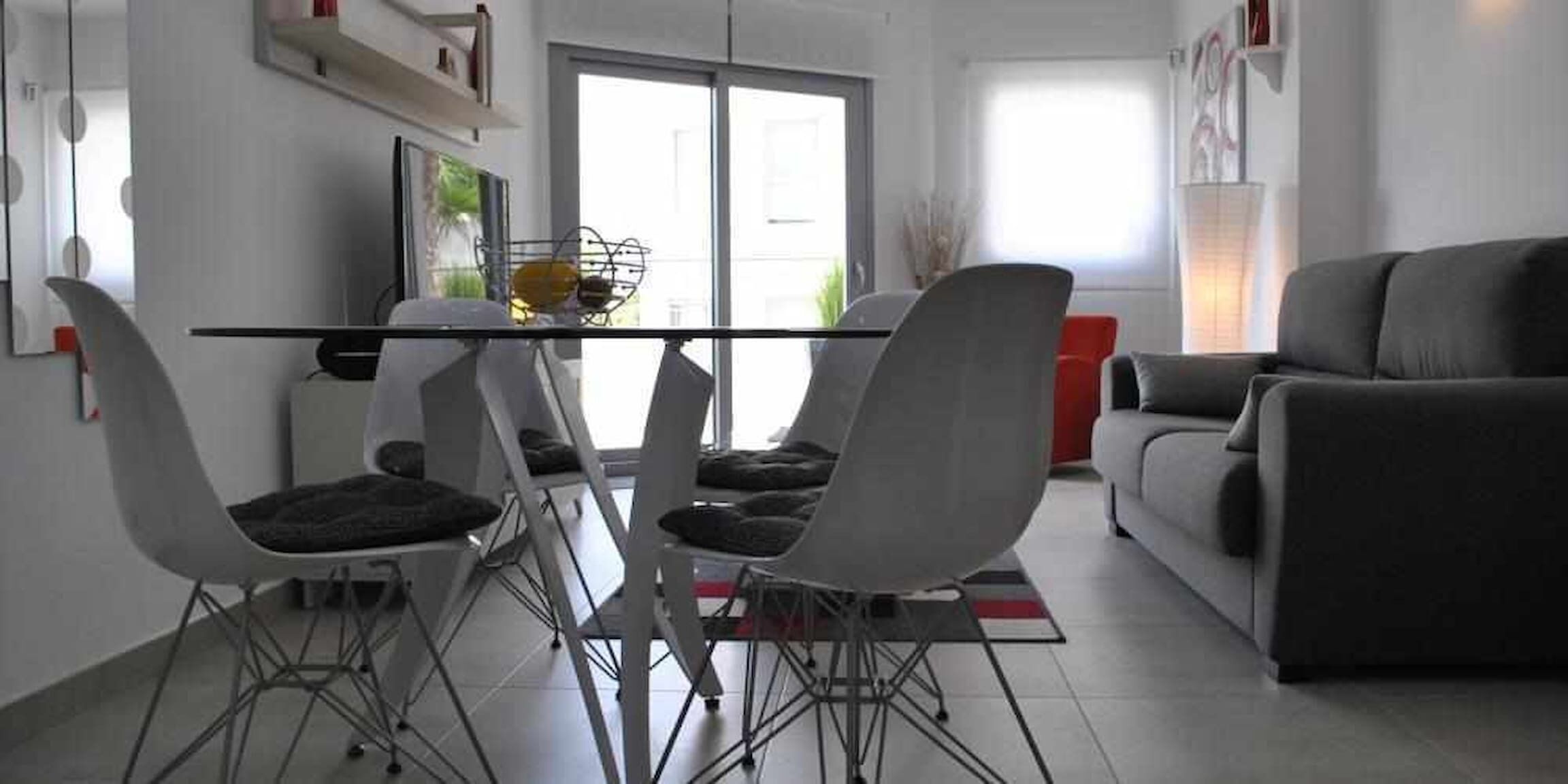 A modern living room with a grey sofa, a dining table with four chairs, and a large window providing natural light.