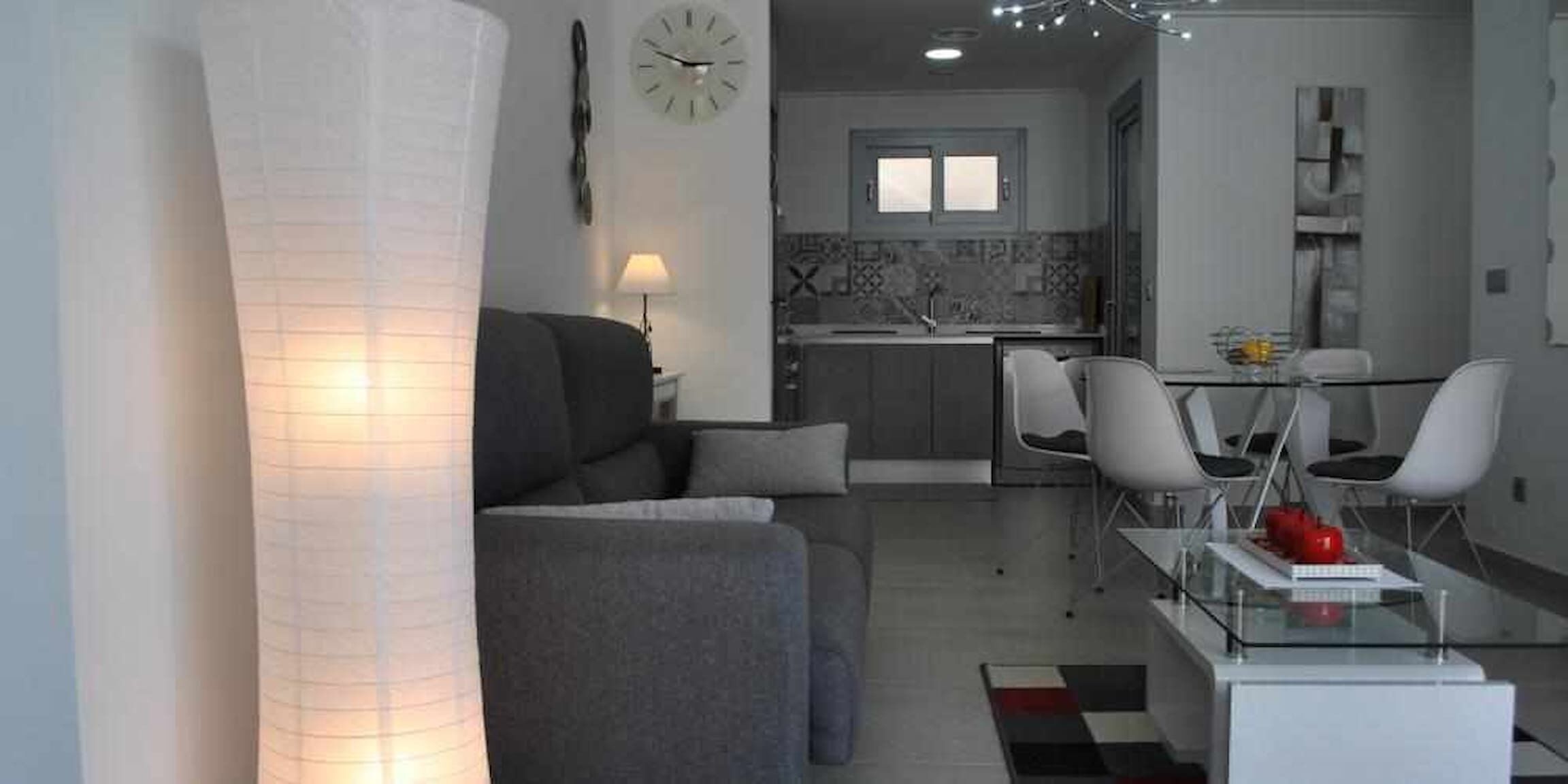 En moderne stue med en sofa, et glasbord og et spiseområde med stole og et lille bord. Et køkken er synligt i baggrunden, med et vindue og et ur på væggen.