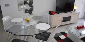 A modern living room with a glass dining table, white chairs, a flat-screen TV, a red speaker, and a small table with apples. - Groundfloor Property Amalia Torrevieja (Torrevieja)