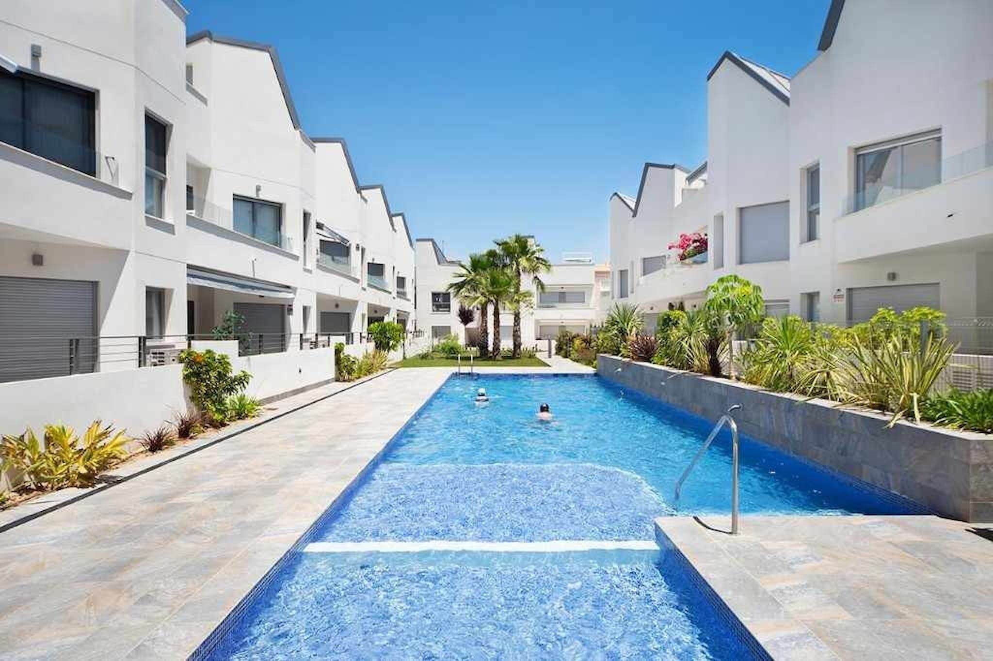 A modern hotel with a central swimming pool, surrounded by white buildings and palm trees.