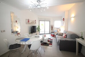 A modern living room with a dining area, a grey sofa, a TV, and a large window. - Groundfloor Property Amalia Torrevieja (Torrevieja)
