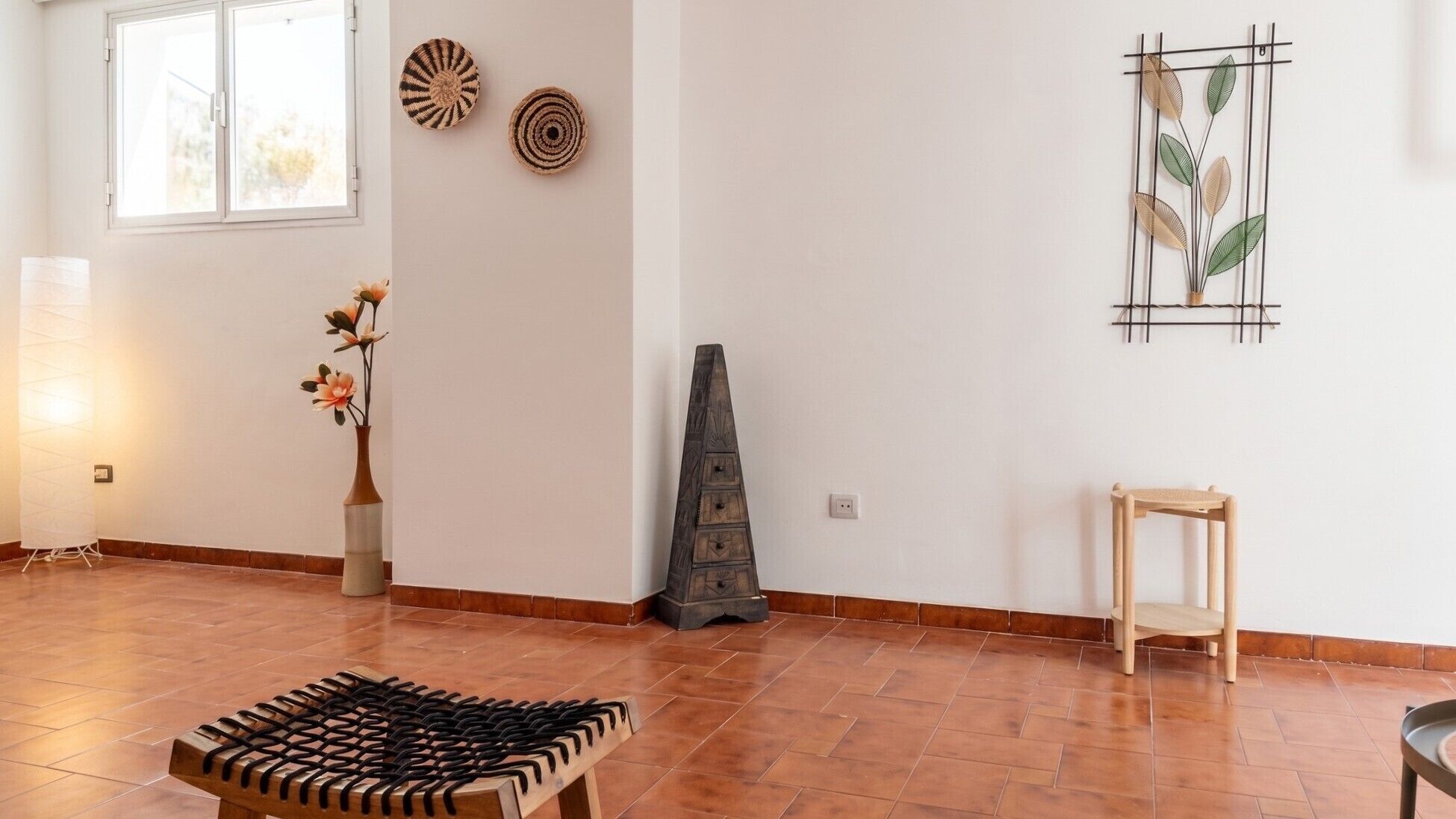 A room with a tiled floor, a wooden bench, a vase with flowers, a wall-mounted shelf with decorative items, and a window with blinds.