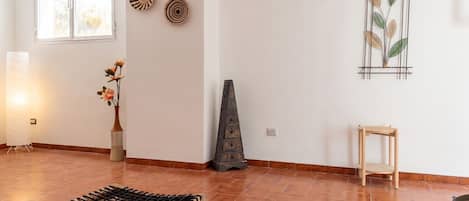 A room with a tiled floor, a wooden bench, a vase with flowers, a wall-mounted shelf with decorative items, and a window with blinds.