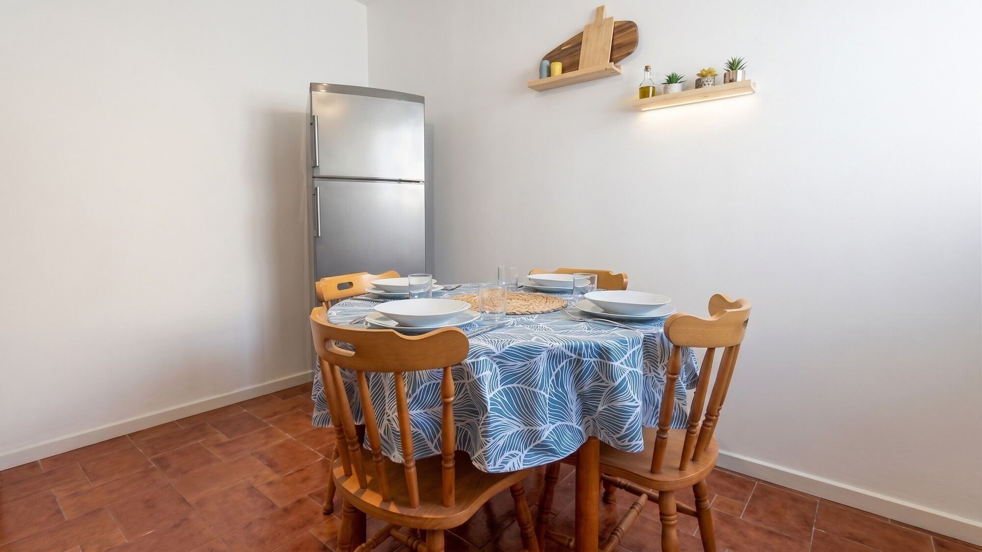 A dining area with a table set for four, wooden chairs, a patterned tablecloth, and a modern refrigerator.