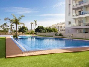 A swimming pool with a wooden deck, surrounded by green grass and palm trees. - Apartment Aqua Nature La Mata (Torrevieja)