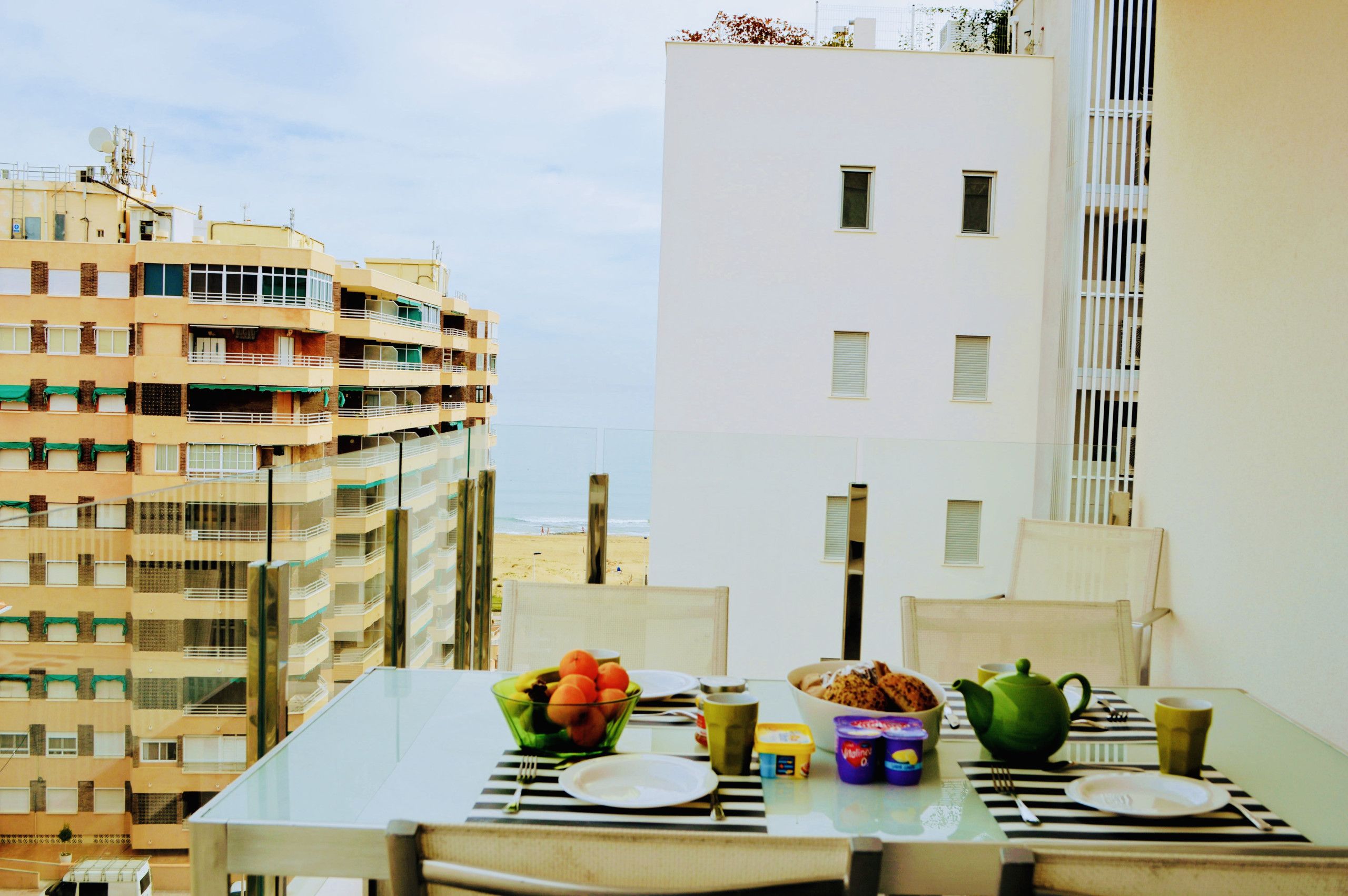 A dining table with a bowl of fruit, a teapot, and a cup, set against a backdrop of a cityscape with buildings and a beach view.