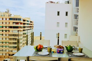 A dining table with a bowl of fruit, a teapot, and a cup, set against a backdrop of a cityscape with buildings and a beach view. - Apartment Aqua Nature La Mata (Torrevieja)
