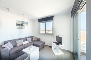 Ein modernes Wohnzimmer mit einem grauen Ecksofa, einem weißen Couchtisch, einem Flachbildfernseher und einem großen Fenster mit Blick auf Gebäude.