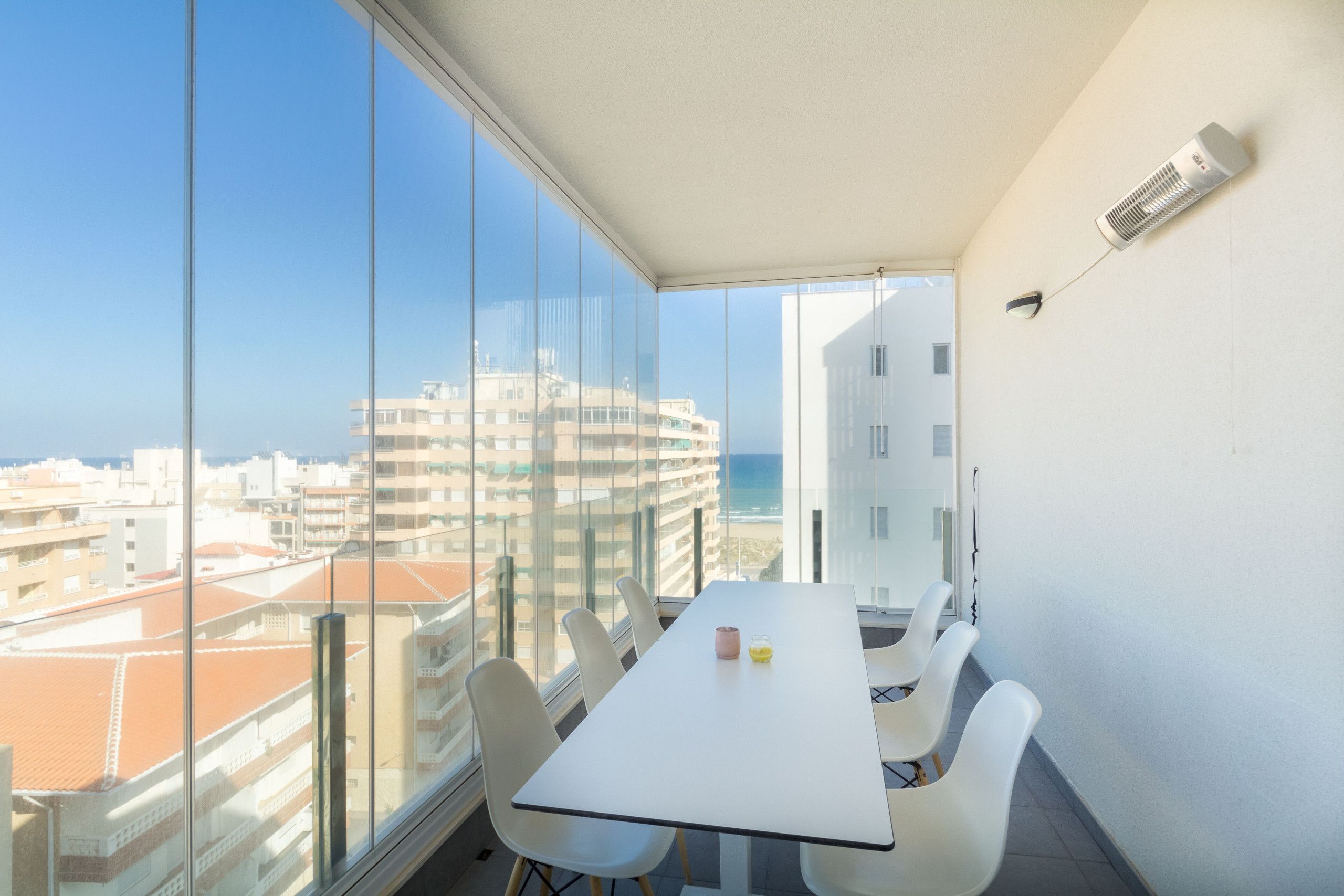 Un balcón con una mesa y sillas blancas, un muro de cristal y vistas a edificios y al mar.
