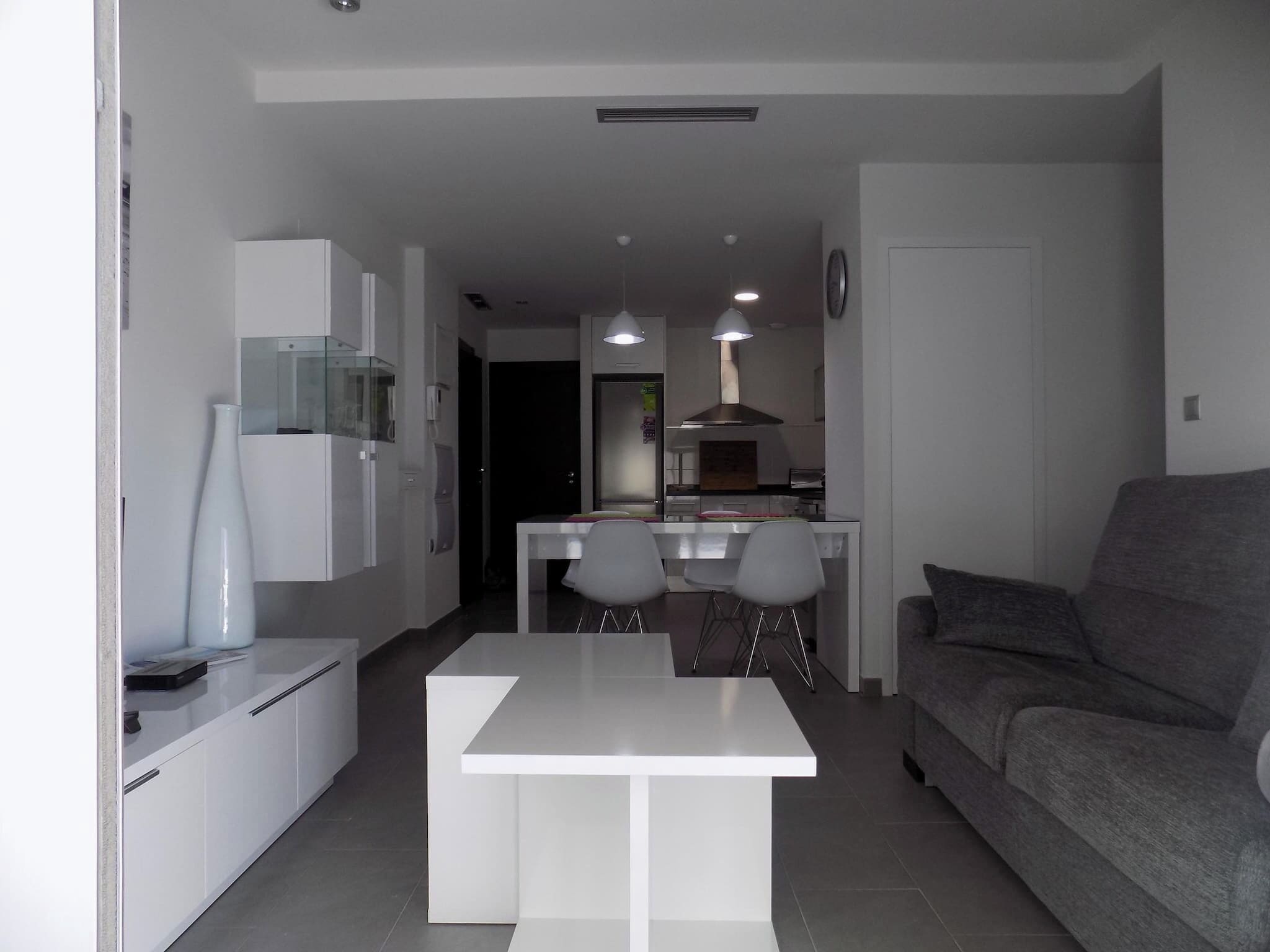 A modern kitchen with white cabinets, a dining table with four chairs, and a grey sofa.