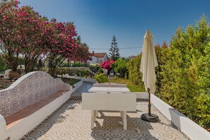 Eine Terrasse mit weißer Bank, Tisch und Sonnenschirm, umgeben von Grünpflanzen und einem gemusterten Boden.