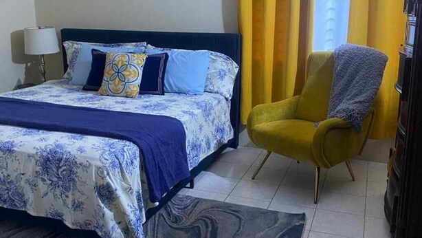 A bedroom with a bed, a yellow armchair, a lamp, and a painting on the wall. - Havendale Suite Retreat (Kingston)