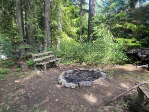 Ein bewaldetes Gelände mit Feuerstelle, einer Holzbank und einem Holzstapel.