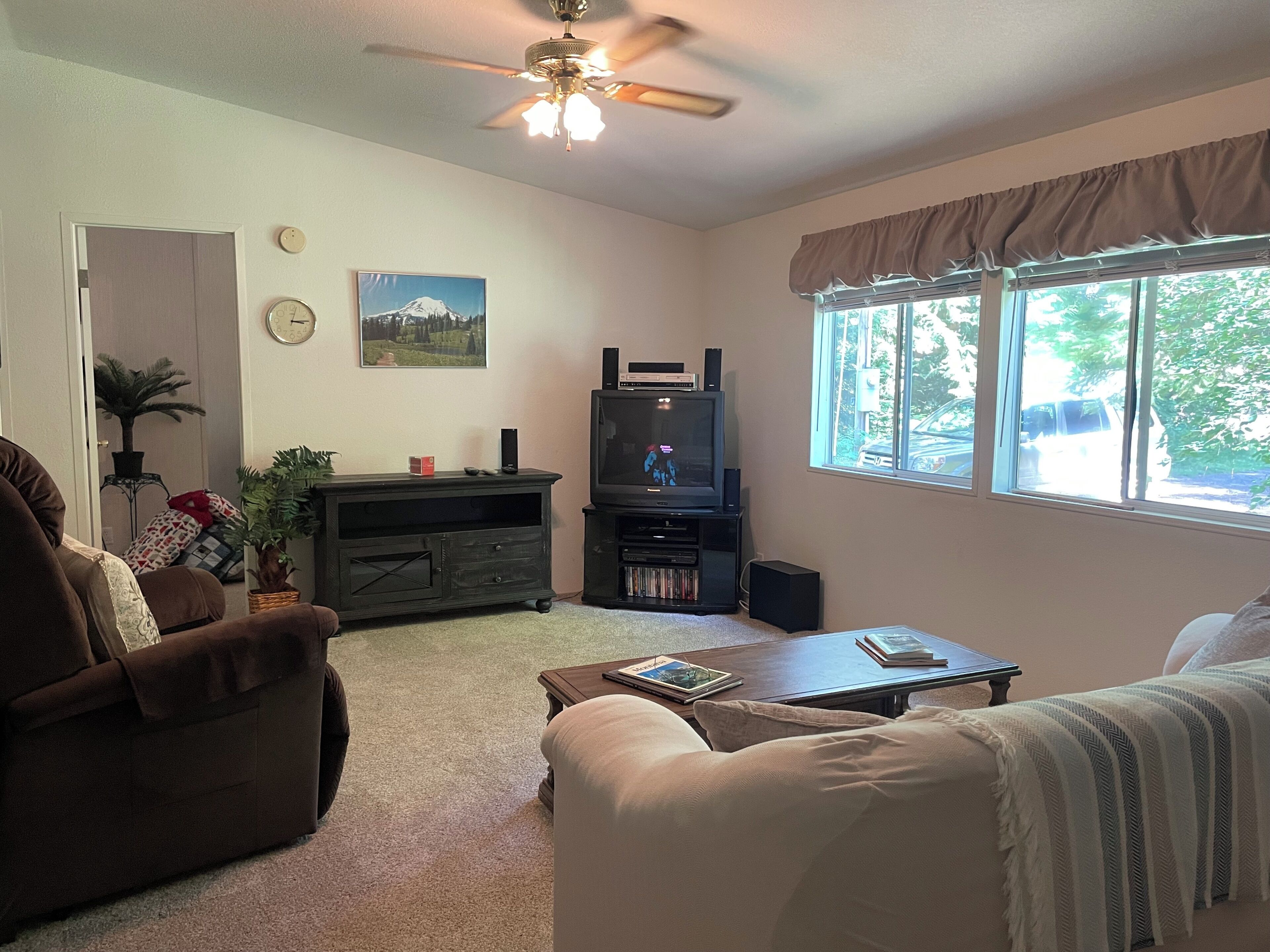 A living room with a sofa, a coffee table, a TV stand with a television, a ceiling fan, and a window with curtains.