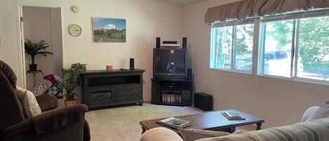 A living room with a sofa, a coffee table, a TV stand with a television, a ceiling fan, and a window with curtains.