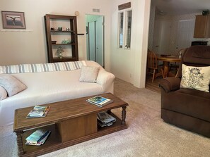 Ein Wohnzimmer mit einer Couch, einem Couchtisch, einem braunen Sessel, einem Bücherregal aus Holz und einem Essbereich im Hintergrund.