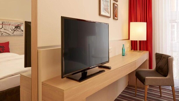 A hotel room with a flat-screen TV on a wooden console, a bed with white linens, a red curtain, and a striped carpet. - 2 Person Holiday Park Home in Stade (Stade)