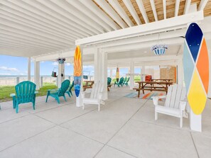 A covered outdoor seating area with chairs and tables, a surfboard decoration, and a view of the beach. - Beachfront, Hot Tub, Arcade, Foosball, Steps to beach, outdoor games & MORE (Galveston)