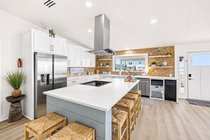 A modern kitchen with a central island, white cabinetry, and a 'WELCOME' sign. - Beachfront, Hot Tub, Arcade, Foosball, Steps to beach, outdoor games & MORE (Galveston)