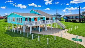A turquoise building with a balcony, a covered patio area, and a sandy ground. - Beachfront, Hot Tub, Arcade, Foosball, Steps to beach, outdoor games & MORE (Galveston)