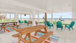 Een overdekt terras met houten tafels en stoelen, uitzicht op het strand en een surfplank.