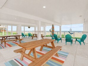 A covered outdoor seating area with wooden tables and chairs, a view of the beach, and a surfboard. - Beachfront, Hot Tub, Arcade, Foosball, Steps to beach, outdoor games & MORE (Galveston)