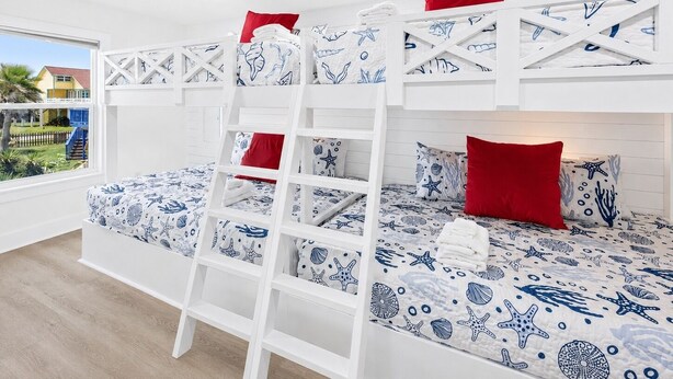 Een slaapkamer met stapelbed, een ladder, witte muren en uitzicht op een huis en groen buiten.