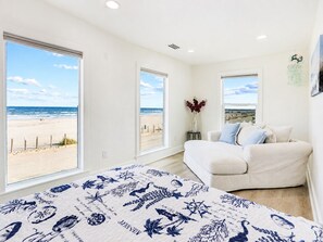 A bedroom with a large window overlooking a beach, a bed with a nautical-themed bedspread, and a white sofa with a vase of flowers. - Beachfront, Hot Tub, Arcade, Foosball, Steps to beach, outdoor games & MORE (Galveston)