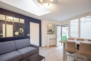 Ein modernes Wohnzimmer mit einer Couch, einem Esstisch mit Stühlen, einer Küchenzeile und großen Fenstern.