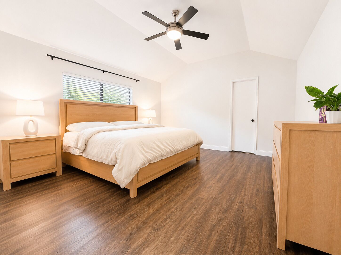A bedroom with a bed, bedside tables, a ceiling fan, a window with blinds, a door, and a plant.