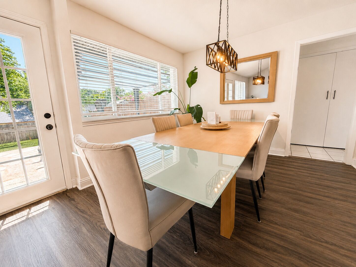 Een eetgedeelte met een tafel met glazen blad, beige stoelen, een houten vloer en een hanglamp.