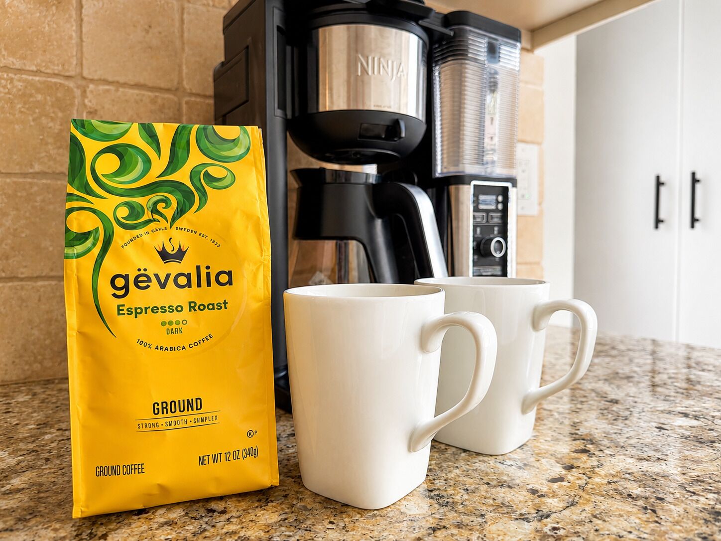 A coffee maker, a bag of coffee, and two white mugs on a kitchen counter.