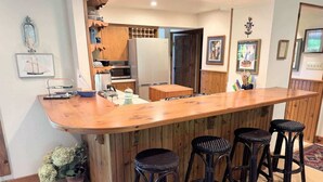 A kitchen with wooden cabinets, a microwave, a refrigerator, a wooden bar counter with stools, and a potted plant. - Island Nights Classic Beach Cottage | Private Association Beach Access (South Haven)