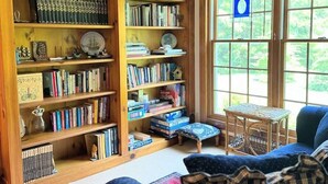 A cozy living room with a large wooden bookshelf filled with books and decorative items, a blue sofa with plaid cushions, and a small table. - Island Nights Classic Beach Cottage | Private Association Beach Access (South Haven)